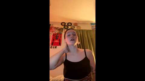 Video of a woman with light skin and auburn hair, wearing a black tank top, standing in a cozy room with a green curtain, red posters, and a warm, dimly lit atmosphere.