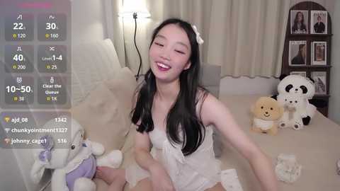 Video of an Asian woman with long black hair, wearing a white lace lingerie set, smiling in a bedroom with stuffed animals, and a digital overlay of a dating app screen.