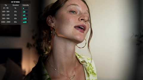 A video of a fair-skinned, middle-aged woman with brown hair tied back, wearing a green floral blouse, standing in a dimly lit room with a blurred background.