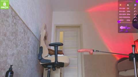 Video of a narrow, dimly lit room with a beige chair, a wooden desk, and a computer monitor emitting a red light. The walls are plain white, and the door is slightly ajar.