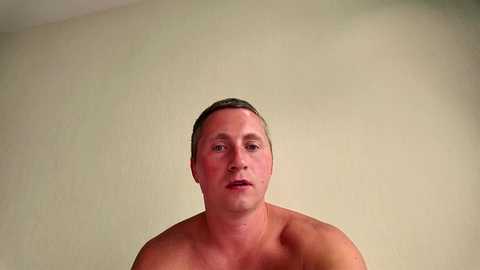 Video of a shirtless, middle-aged Caucasian man with short brown hair, fair skin, and a neutral expression against a plain beige wall.