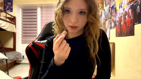 Video of a young blonde girl with blue eyes, wearing a black top, sitting in a red gaming chair, surrounded by colorful posters and a bunk bed in a well-lit room.