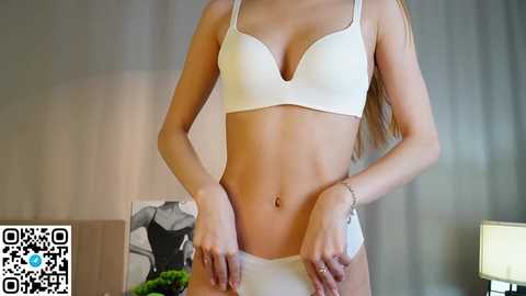 Video of a slender, fair-skinned woman in white lingerie, standing indoors with a blurred, modern room background.