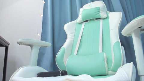 Video of a mint-green gaming chair with white accents and a lumbar support cushion, positioned against a blue curtain backdrop, featuring a minimalist office setting.