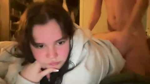 Video of a nude man penetrating a woman from behind on a bed, her expression neutral, wearing a light hoodie, blurred background.