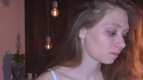A video of a young Caucasian woman with long, straight, light brown hair, wearing a white tank top, gazing introspectively. She has fair skin and a delicate facial structure. Two exposed light bulbs hang in the background, casting a warm, dim glow.