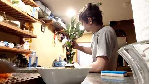 Video of a young Asian man with short brown hair, wearing a gray T-shirt, preparing vegetables on a kitchen counter. The kitchen is cluttered with dishes, utensils, and a water bottle, with wooden shelves and a white ceiling light.