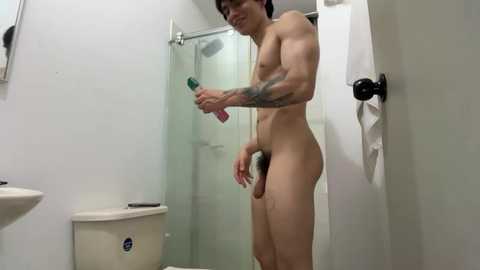 Video of a muscular, tattooed, nude man with a flaccid penis, holding a pink bottle in a white-tiled bathroom with a glass shower, white towels, and a toilet.