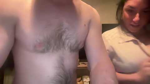 Video of a topless, hairy-chested man standing next to a woman with shoulder-length hair, both indoors.