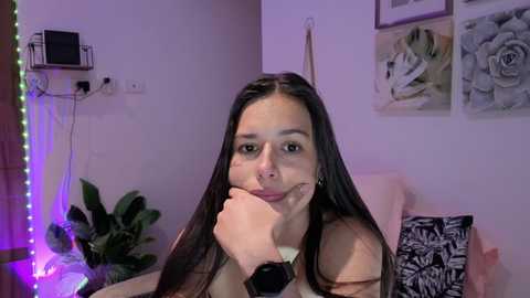 A video of a young woman with long black hair, light skin, and a neutral expression, sitting in a room with purple lighting, plants, and floral artwork on the wall.