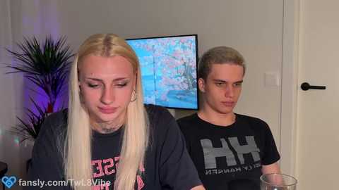 Video of two transgender individuals, a blonde woman and a man with short, dyed hair, wearing black t-shirts. The woman appears distressed, the man looks disinterested. Background includes a flat-screen TV displaying a nature scene and a plant.