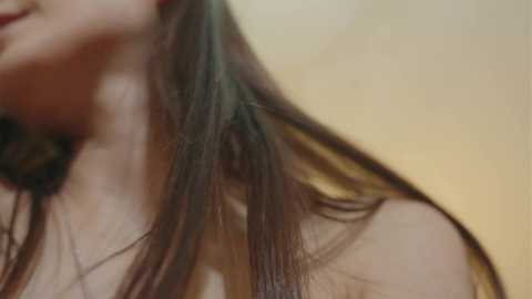 A close-up video of a young woman's head and shoulder, showing her long, dark brown hair cascading down, with a blurred beige background. Her lips are slightly parted, revealing a hint of a smile. The image is intimate and soft-focus, emphasizing texture and natural beauty.